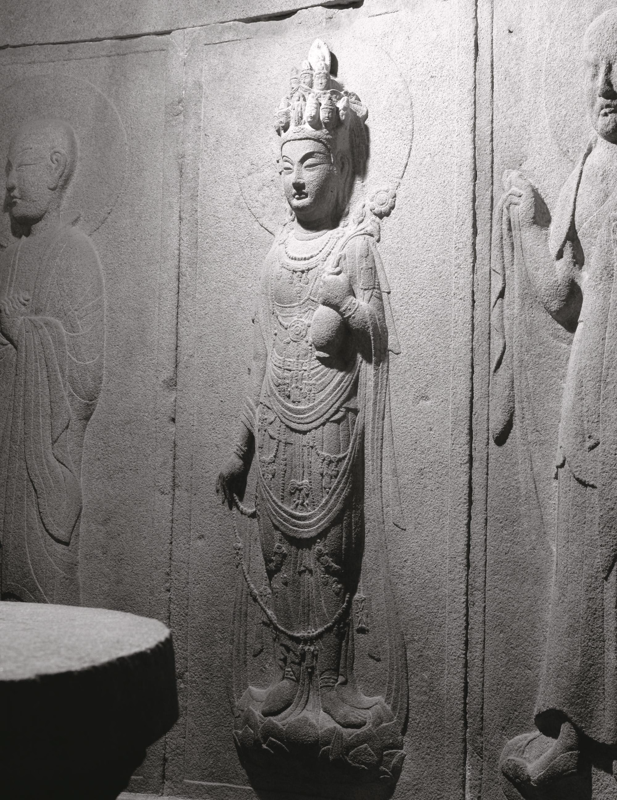 A stone relief carving on a grotto wall depicting the standing Eleven-Headed Avalokiteshvara with a halo and multiple small heads arranged on the crown.
